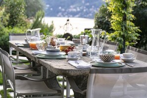 Ein Steintisch, gedeckt für das Frühstück mit verschiedenen Gerichten, Gläsern und einer Karaffe, umgeben von Stühlen.
