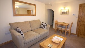 A living room with a beige sofa, a wooden coffee table, and a dining table with chairs. - Saltburn Holidays Primrose Cottage (Guisborough)