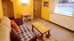 A living room with a sofa, a coffee table, a dining table with chairs, a wooden door, and a window with curtains. - Saltburn Holidays Primrose Cottage (Guisborough)