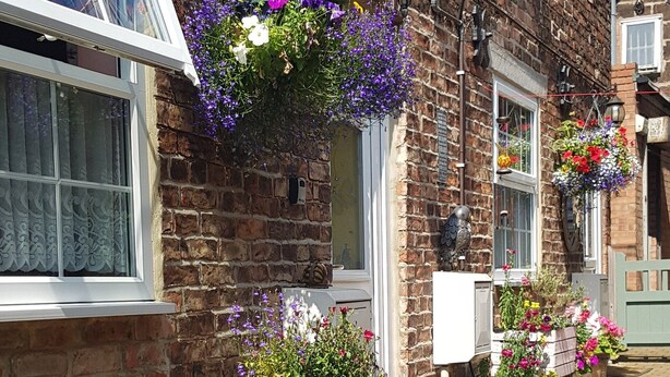 A brick building with hanging flower baskets and a wooden stool with a potted plant. - Saltburn Holidays Primrose Cottage (Guisborough)