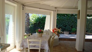 Une terrasse couverte avec une table à manger pour quatre personnes, entourée de chaises. Des plantes en pot, ornées de fleurs, sont disposées sur la table, et de l’extérieur, on peut admirer un paysage verdoyant.