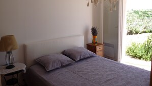 A bedroom with a bed, a bedside table with a lamp, a small wooden nightstand, and a window with a view of greenery.