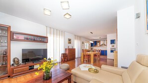 En rummelig opholdsstue med et træbord, en beige sofa og et fjernsyn på et træstel. I baggrunden kan man se et spiseområde med et blåt bord og stole.