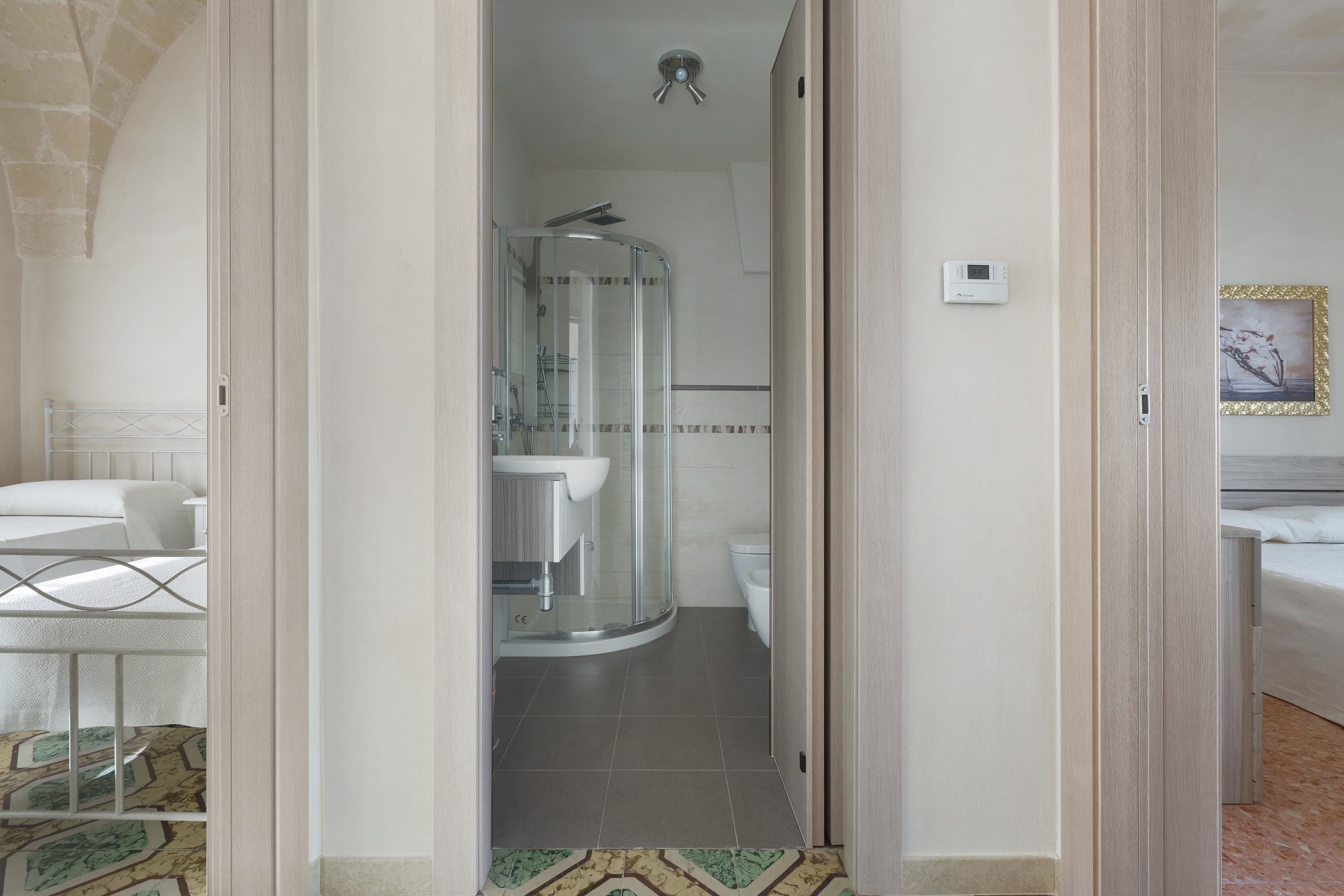 Habitación de hotel con ducha de vidrio, lavamanos blanco y cama con estructura metálica.