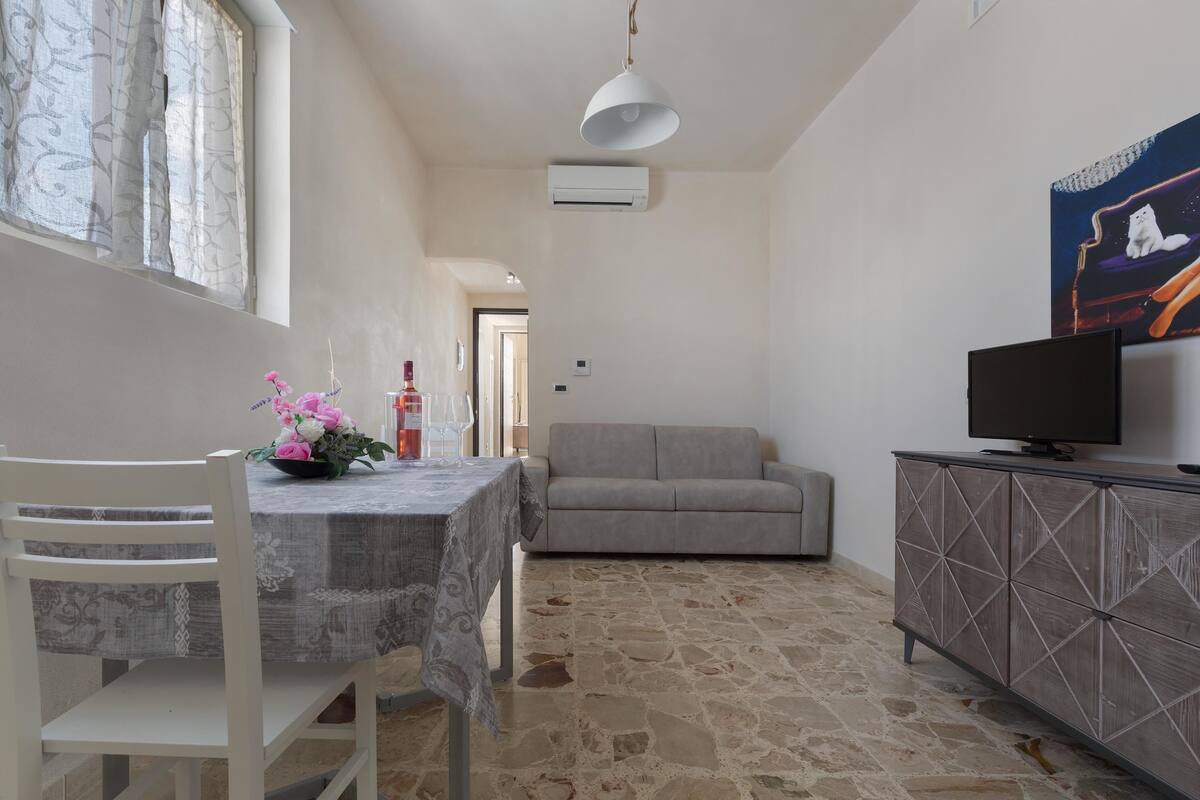 A living room with a sofa, a dining table with chairs, a TV on a cabinet, and a painting on the wall.