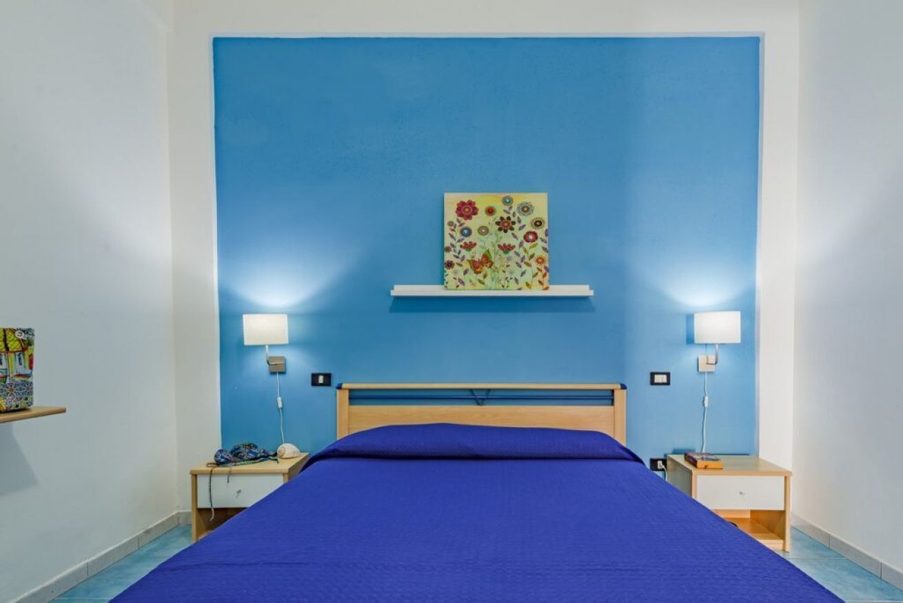 Ein Schlafzimmer mit blauer Wand, einem Bett mit blauer Bettdecke, zwei Nachttischen mit Lampen und einem Blumenbild an der Wand.