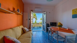 Una stanza con divano, tavolo da pranzo e TV. Una porta dà accesso a una zona esterna con vista su palme e piscina.