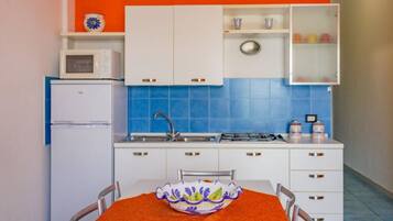 A compact kitchen with white cabinets, a blue tiled backsplash, a microwave, a refrigerator, and a dining table with chairs.