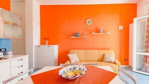 Una stanza con una parete arancione vivace, un divano color beige, un tavolo bianco con una ciotola e un armadietto bianco.