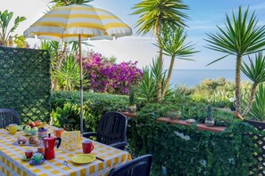A table set outdoors with a checkered tablecloth, a striped umbrella, and various items including a fruit bowl, cups, and a teapot. - Apartment Lumia Superior (Sciacca)