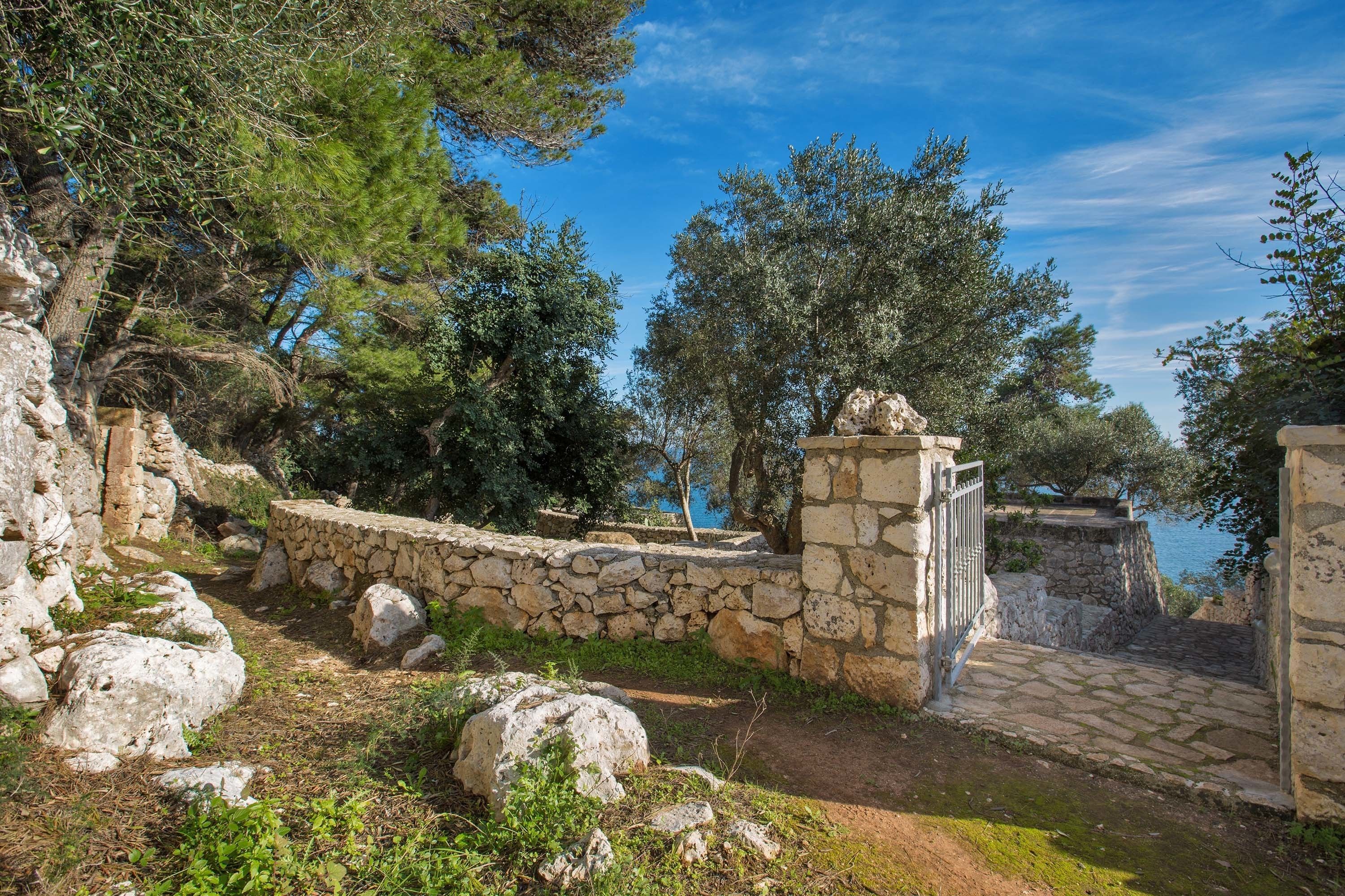 A stone pathway leading to a stone wall with a gate, surrounded by trees and rocky terrain.