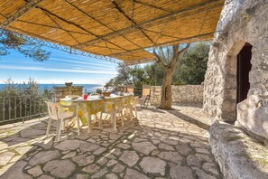 Un espace extérieur aménagé pour les repas, avec un sol en pierre, une table entourée de chaises et une vue sur la mer.