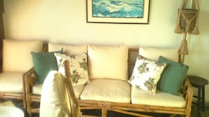 A wicker sofa with floral cushions, a framed painting of a sailboat, and a macrame hanging light. - Village dos Pássaros (Guarapari)