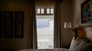 Habitación de hotel con cama, mesita de noche, lámpara y vista al balcón exterior.