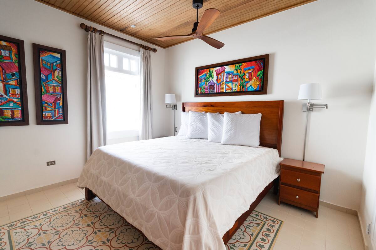 A bedroom with a large bed, a ceiling fan, a nightstand, and two wall-mounted lamps.