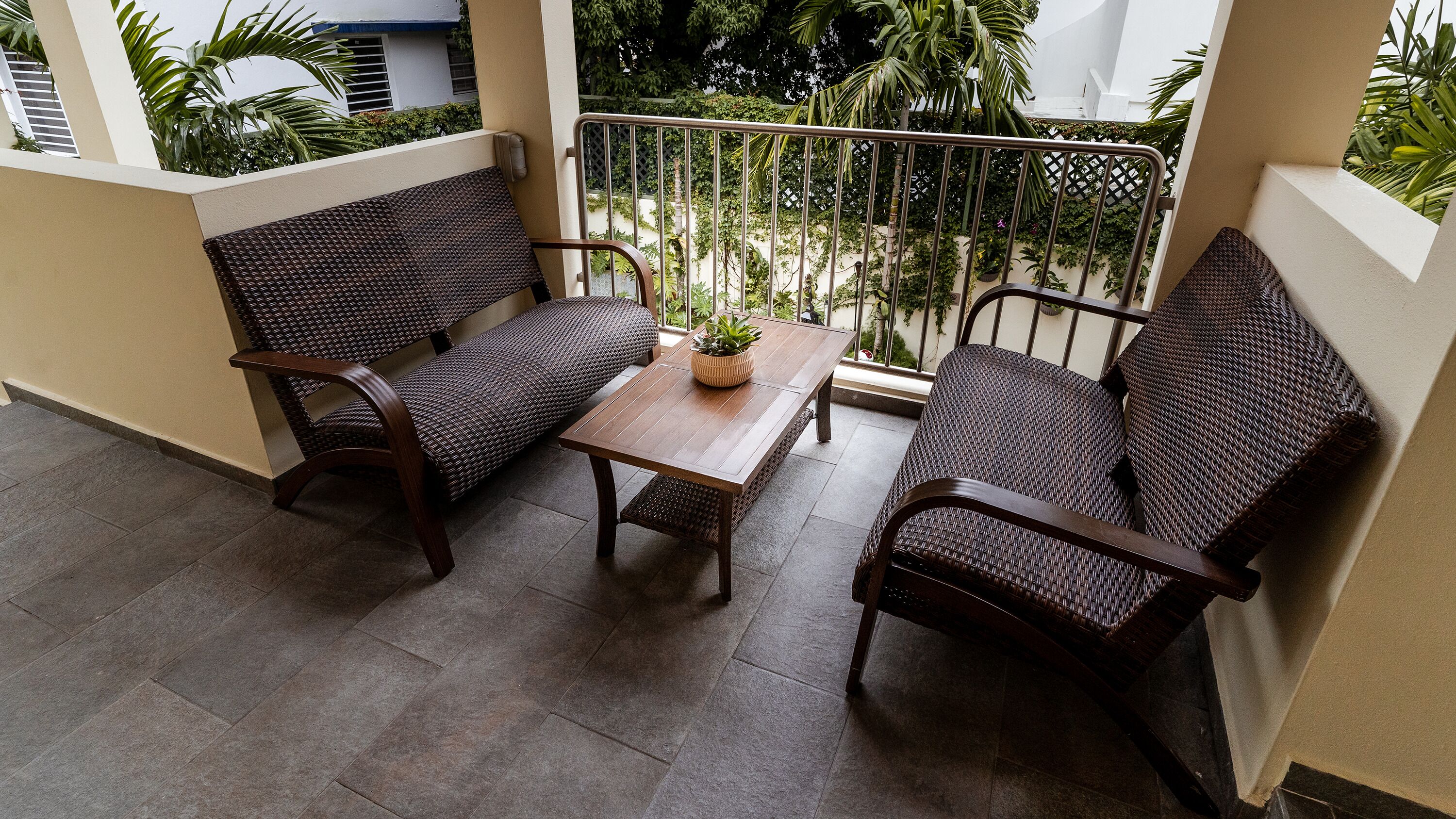 Un balcon agrémenté de deux fauteuils en osier et d’une petite table, entouré d’une rambarde en métal et de plantes en pot.