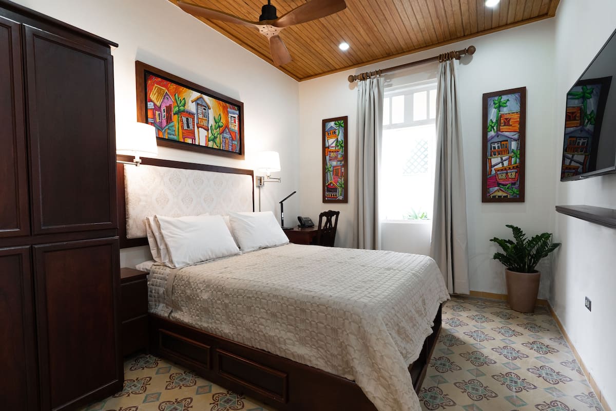 A bedroom with a bed, wooden headboard, a ceiling fan, a nightstand, a lamp, a potted plant, and two wall-mounted paintings.