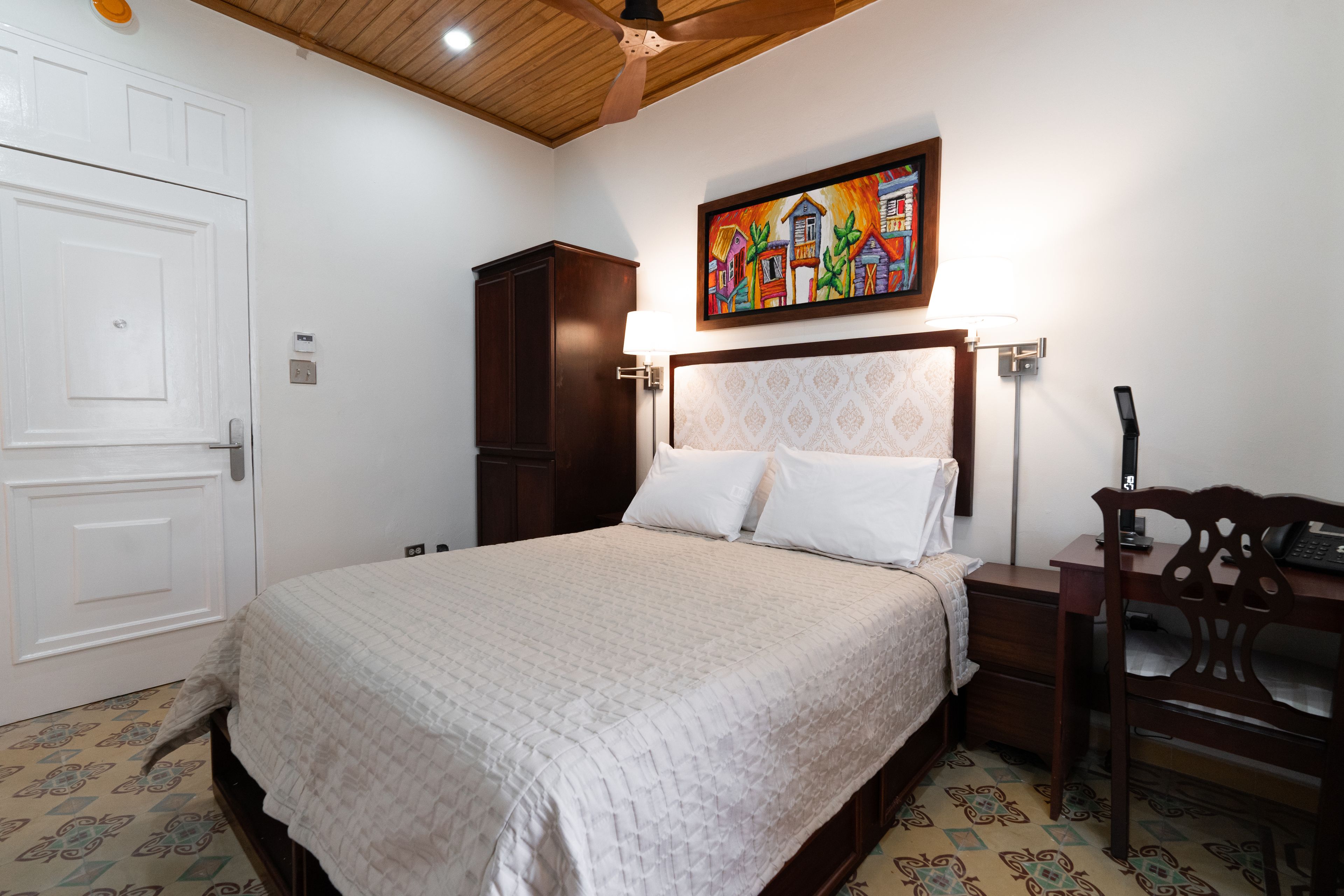 A bedroom with a bed, a desk, a chair, a wardrobe, and a painting on the wall.