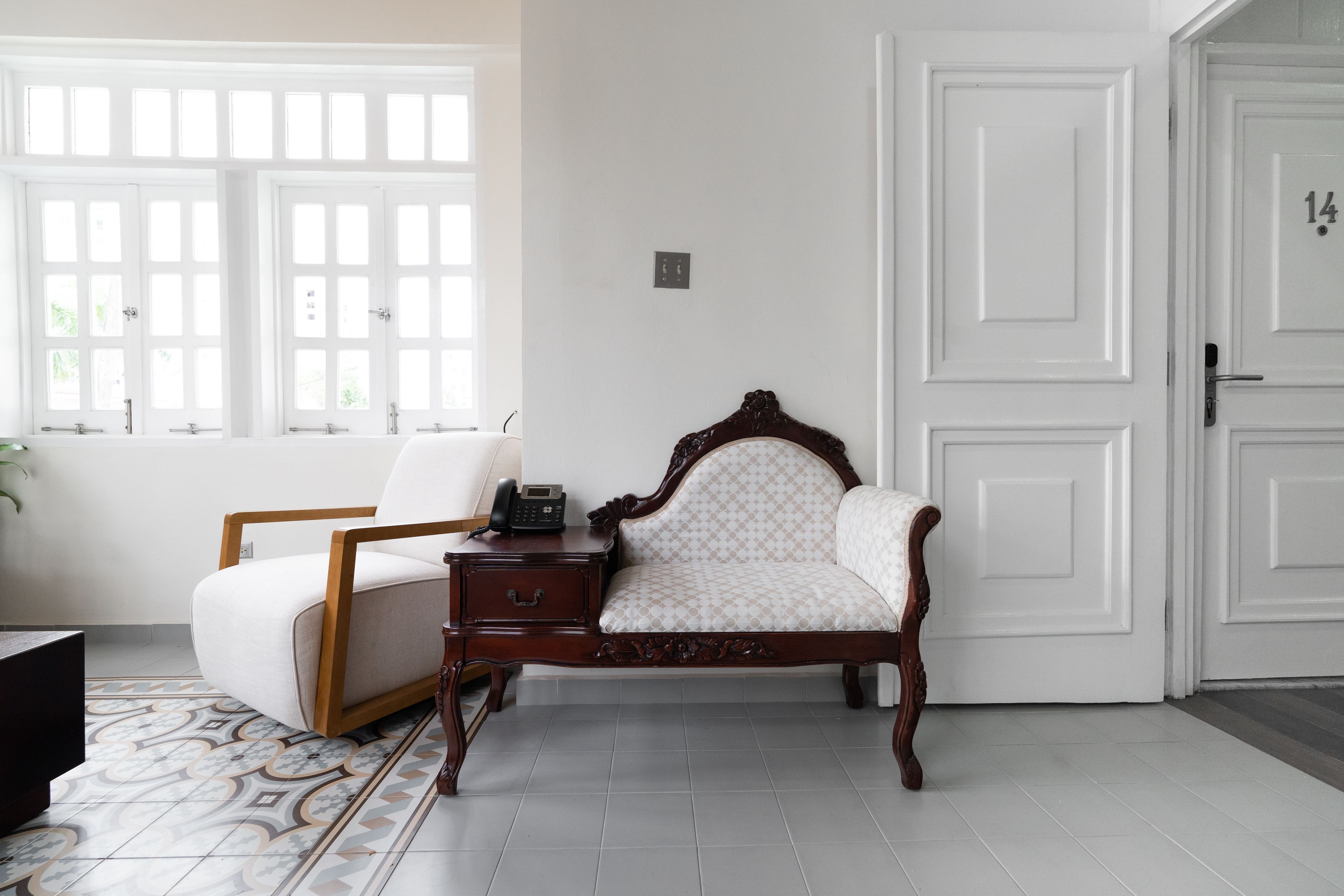 Una habitación con una cama de madera, una silla, una mesita con teléfono y un ventanal con marcos blancos.