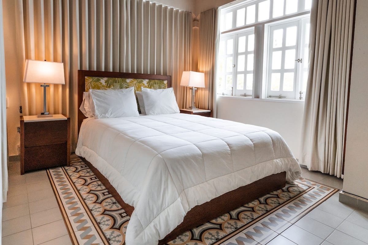 A neatly arranged bedroom with a large bed, two bedside tables with lamps, a patterned rug, and a window with curtains.