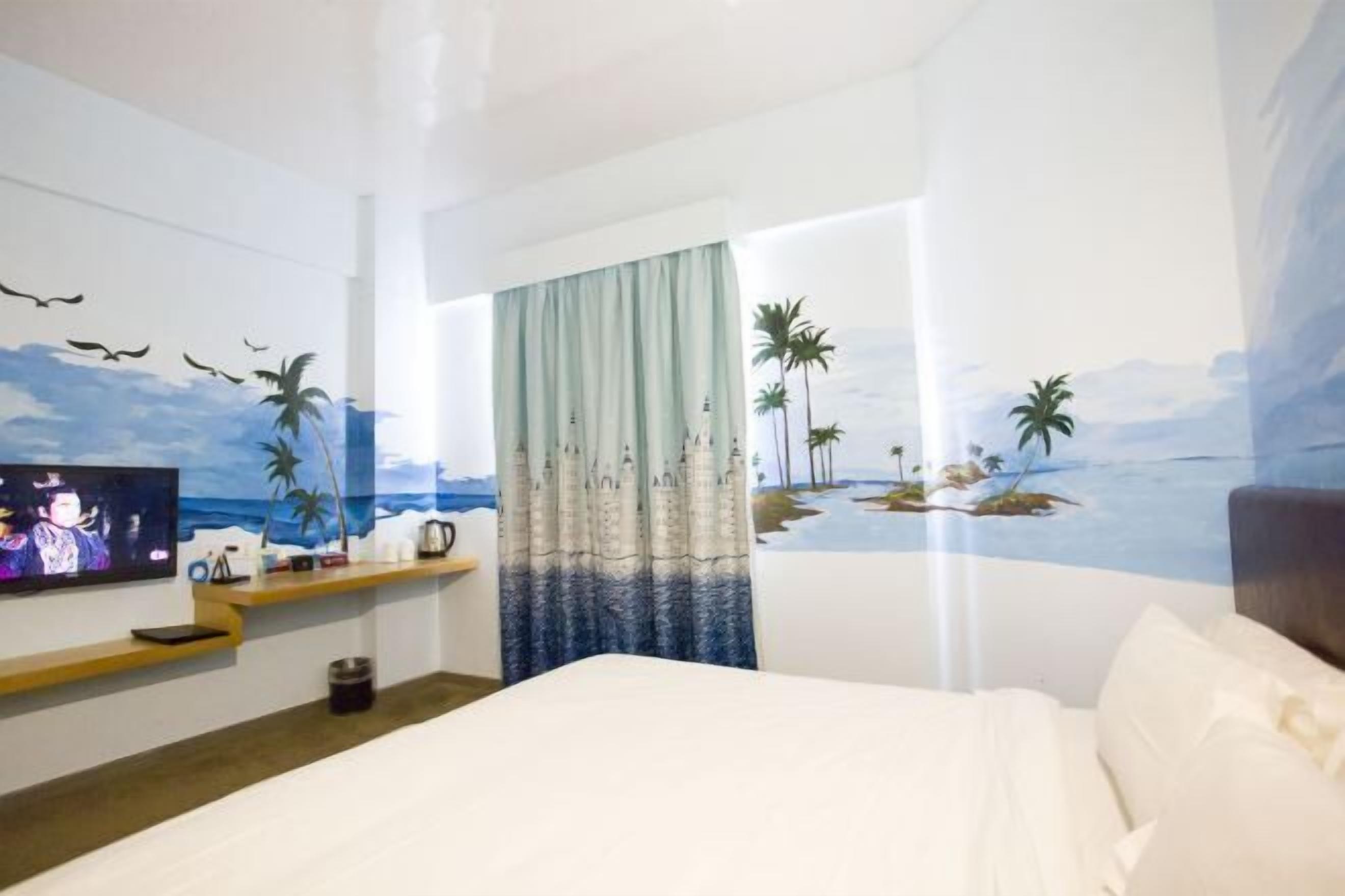 A hotel room with a bed, a TV on the wall, a shelf with items, and a mural of a beach scene.