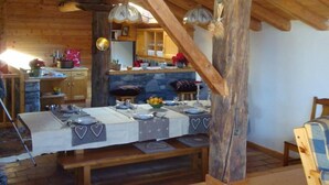 Un intérieur rustique avec des éléments en bois, comprenant une table à manger pour quatre personnes, une cheminée en pierre et une structure de poutres apparentes en bois au plafond.