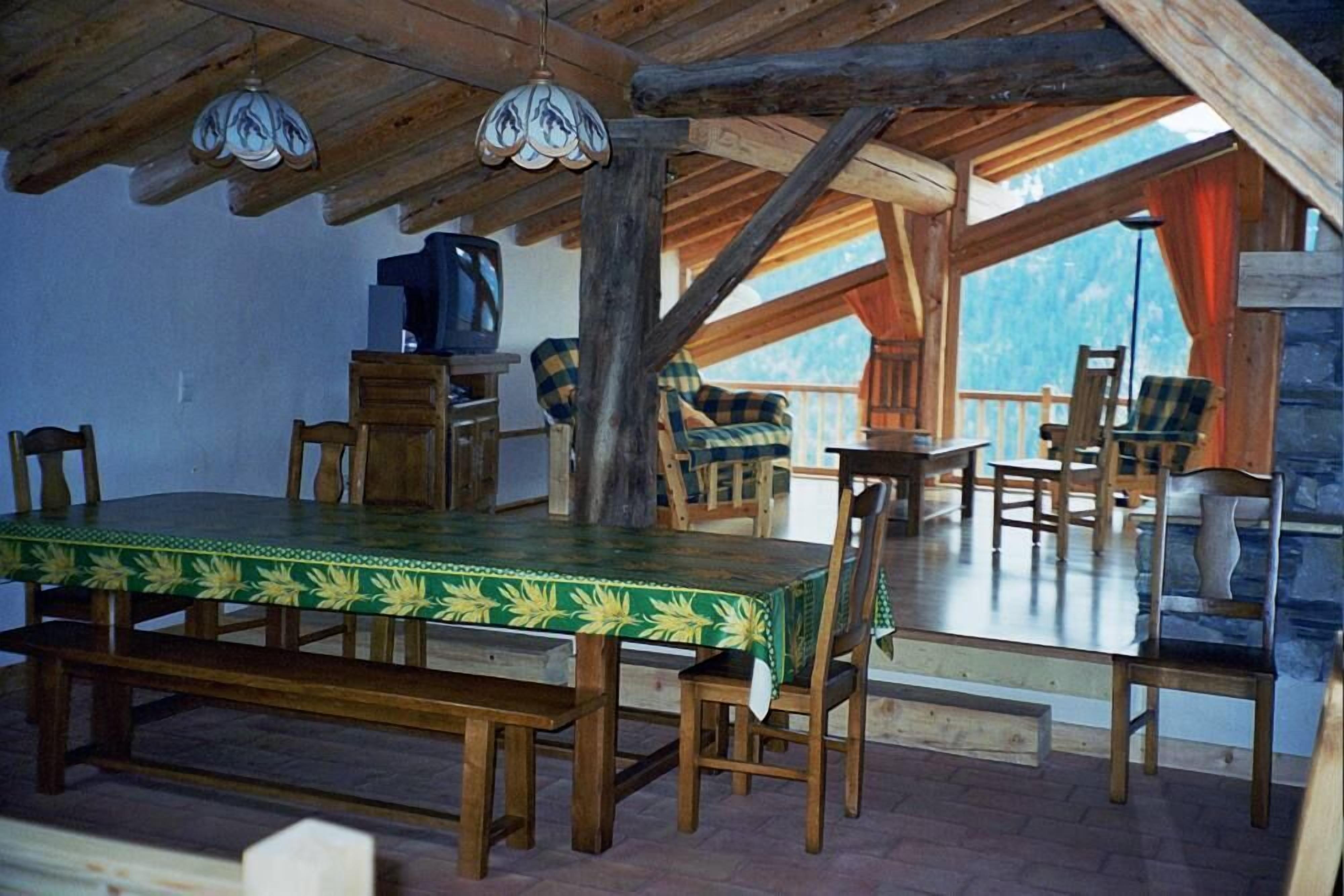 Een rustieke houten eethoek met een tafel, stoelen, een televisie en een balkon met uitzicht op de bergen.
