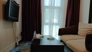 A hotel room with a television mounted on the wall, a sofa, a coffee table with remote controls, and a patterned carpet.