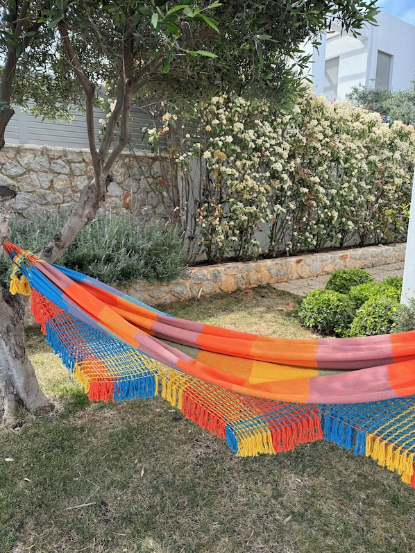 A hammock tied between trees in a garden with a stone wall and flowering bushes in the background.