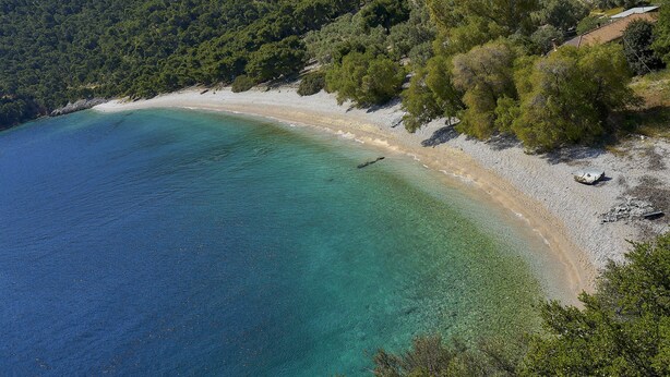 An aerial view of a beach with clear turquoise waters, a sandy shore, and surrounding dense greenery.