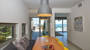 A modern dining area with a wooden table, yellow chairs, and a large grey pendant light. A painting is visible on the wall, and a view of the sea is seen through the glass doors.