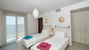 A hotel room with two beds, a large window with a view of the sea, a hanging light fixture, and a wall-mounted fan.