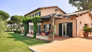 Ein zweistöckiges Haus mit überdachter Terrasse, einem Garten mit Kübelpflanzen und einem gepflasterten Einfahrt.