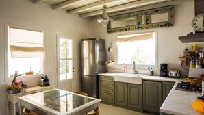 Eine Küche mit Kühlschrank, Spüle und diversen Geräten. Es gibt Fenster mit Vorhängen, einen Deckenventilator und eine Uhr an der Wand.