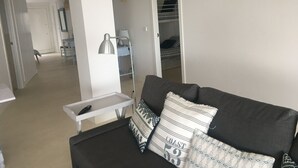 Un salon moderne avec un canapé foncé, une table d’appoint blanche et une lampe. On aperçoit un lit superposé au fond.