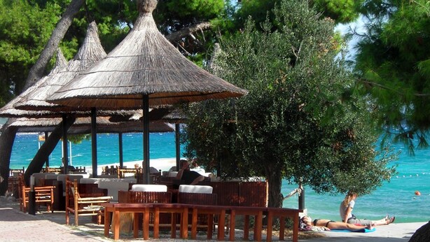 An outdoor seating area with wooden furniture and thatched umbrellas, overlooking a beach and the sea. - New apartments  near the sea   (Seget Donji)