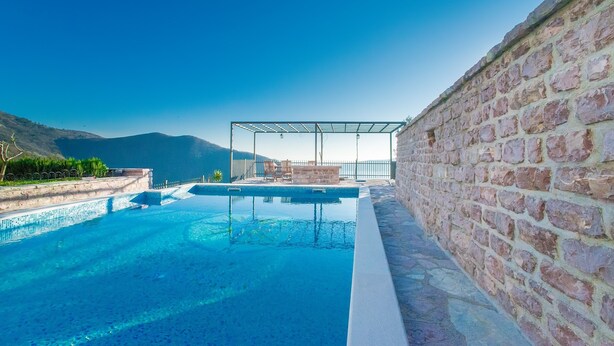 Une piscine offrant une vue dégagée sur les montagnes en arrière-plan, un mur de briques d’un côté et un espace détente couvert.