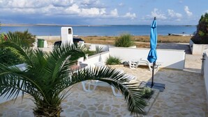 Un espace patio doté d’un sol en pierre, de chaises longues blanches, d’un parasol bleu et offrant une vue sur la mer.