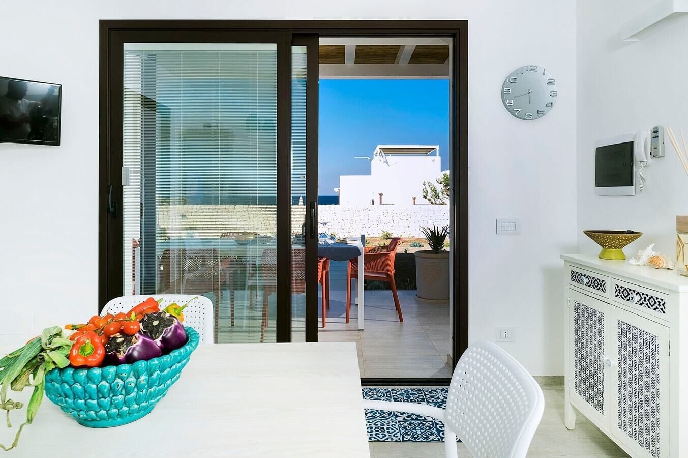A modern living room with a glass door leading to a balcony, a white dining table, and a decorative bowl of fruit.