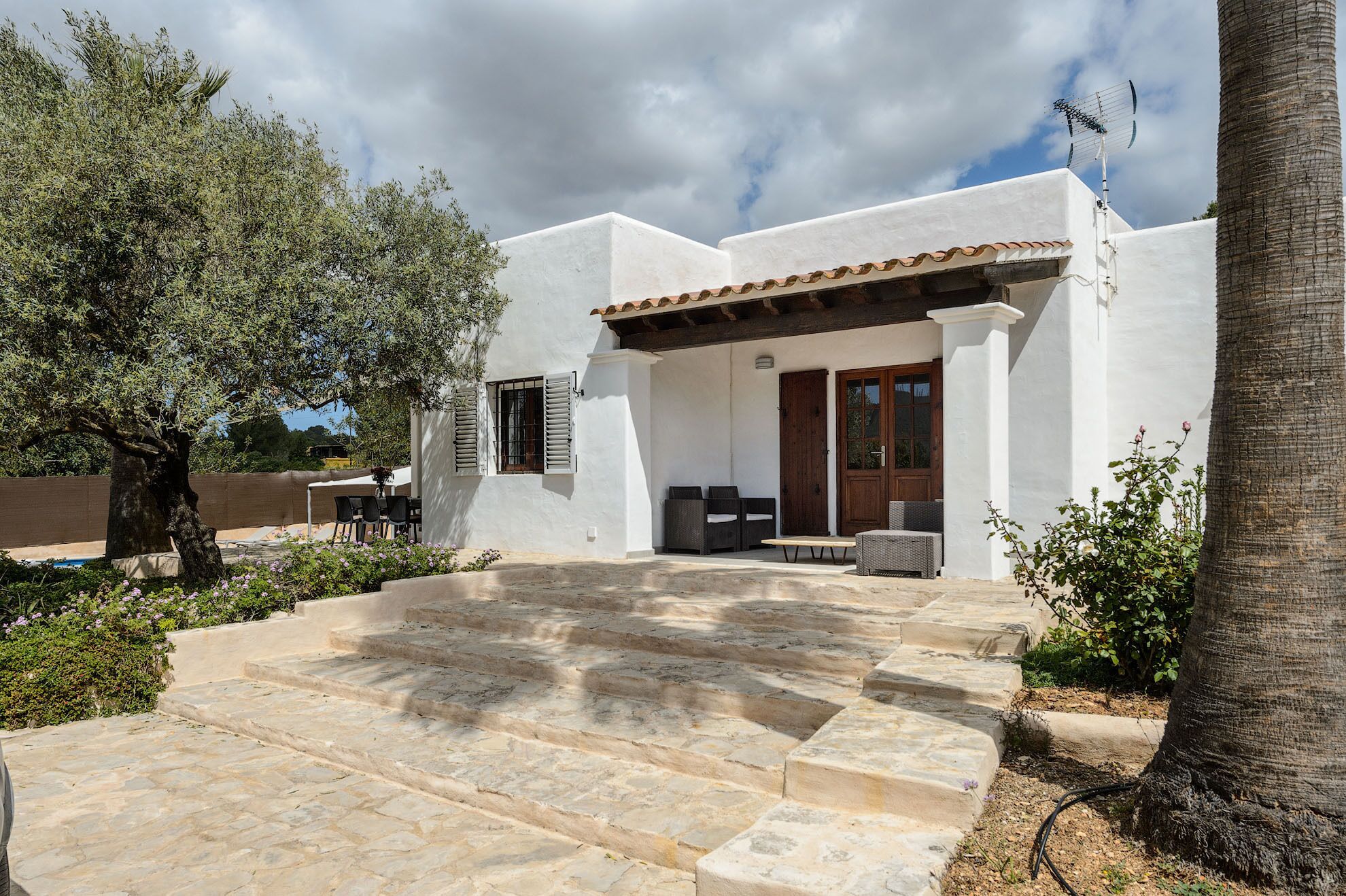 A white building with a stone staircase leading to a wooden door, surrounded by greenery and a palm tree.