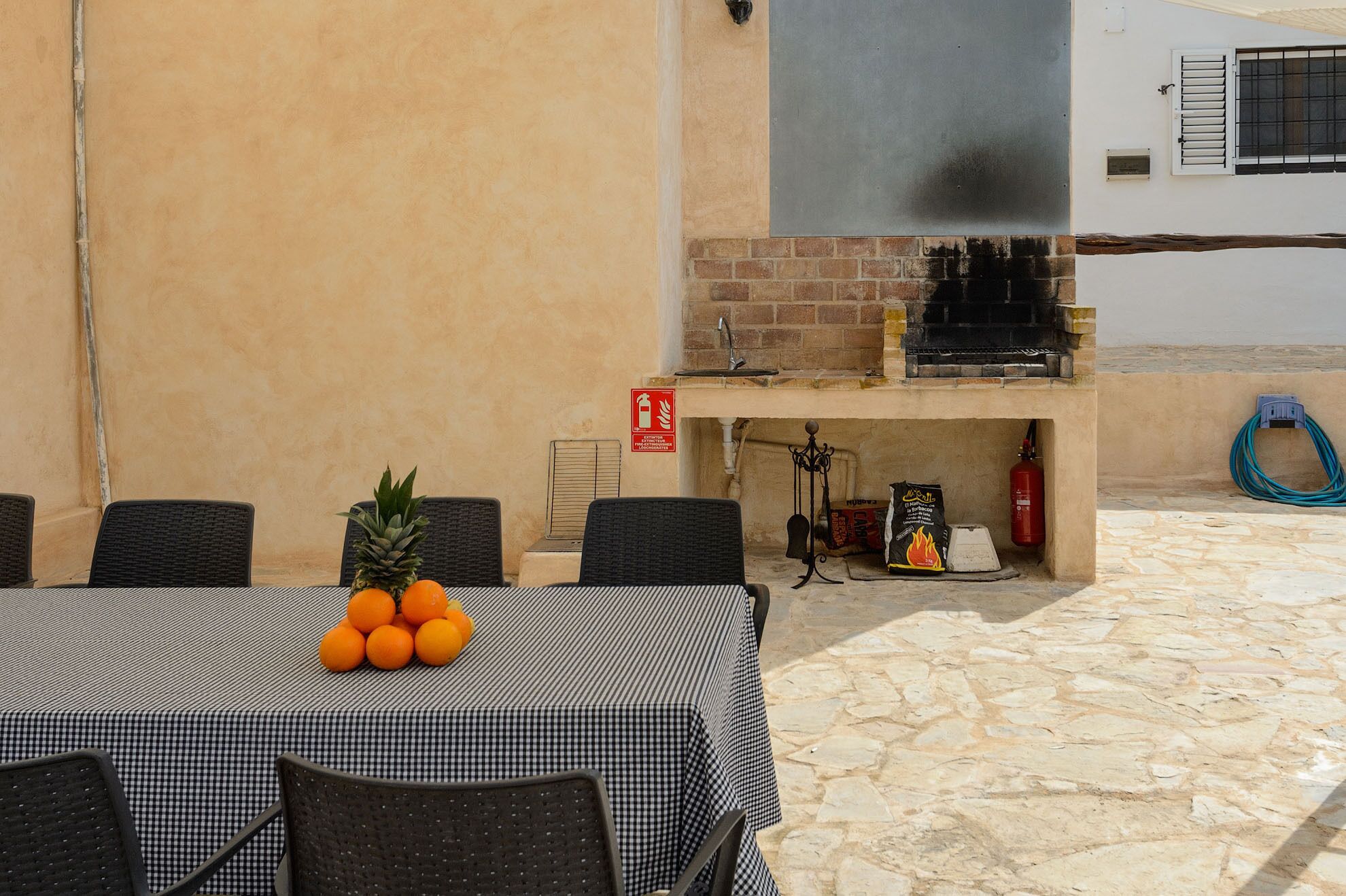 An outdoor dining area with a checkered tablecloth, wicker chairs, a pineapple, and oranges on the table.