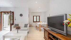 A modern living room with a white sofa, a dining table with chairs, a TV on a wooden stand, and a window with a view of greenery.