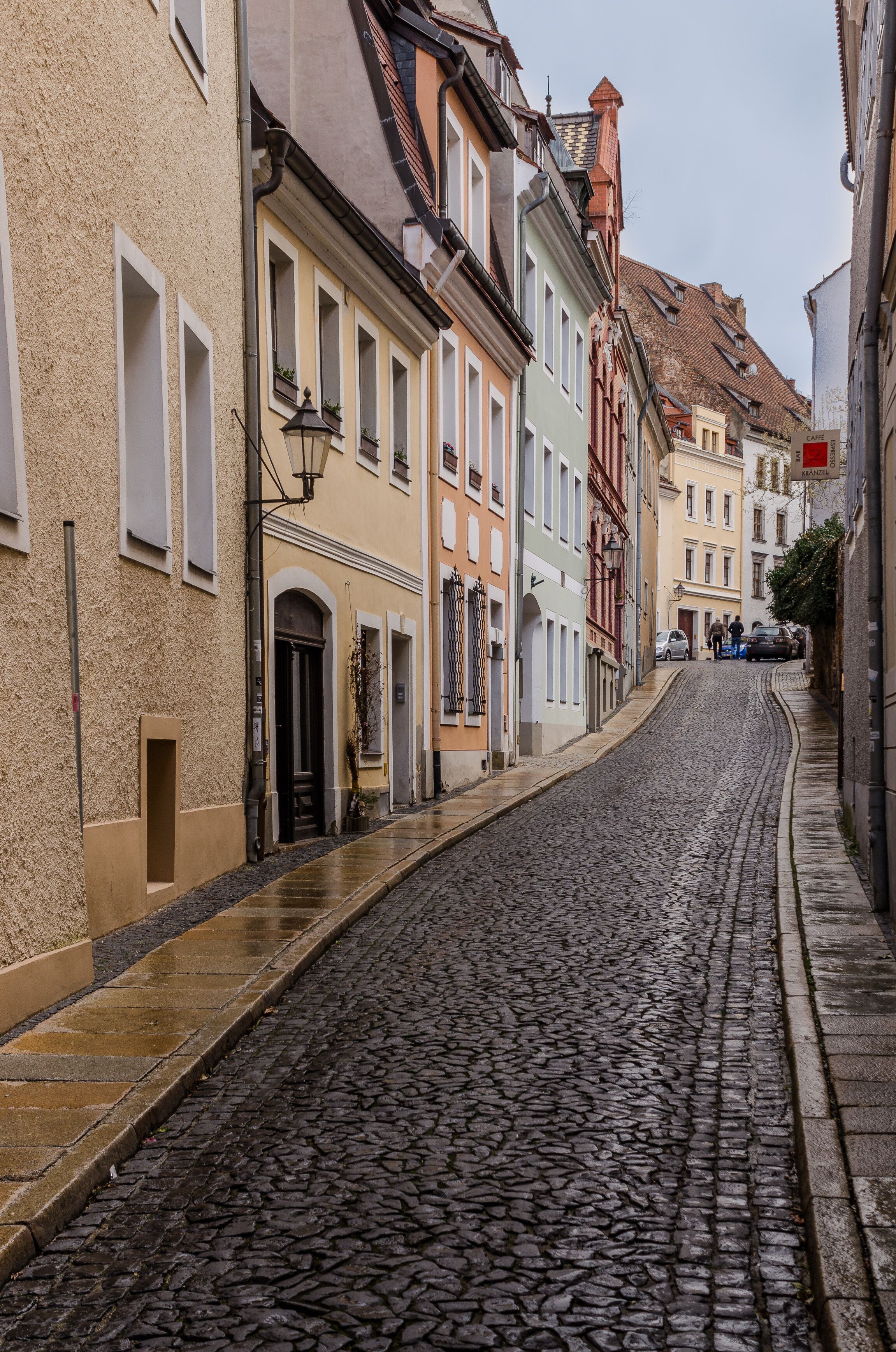 Eine Kopfsteinpflasterstraße gesäumt von bunten Gebäuden, ein schmaler Bürgersteig und einige Fußgänger.