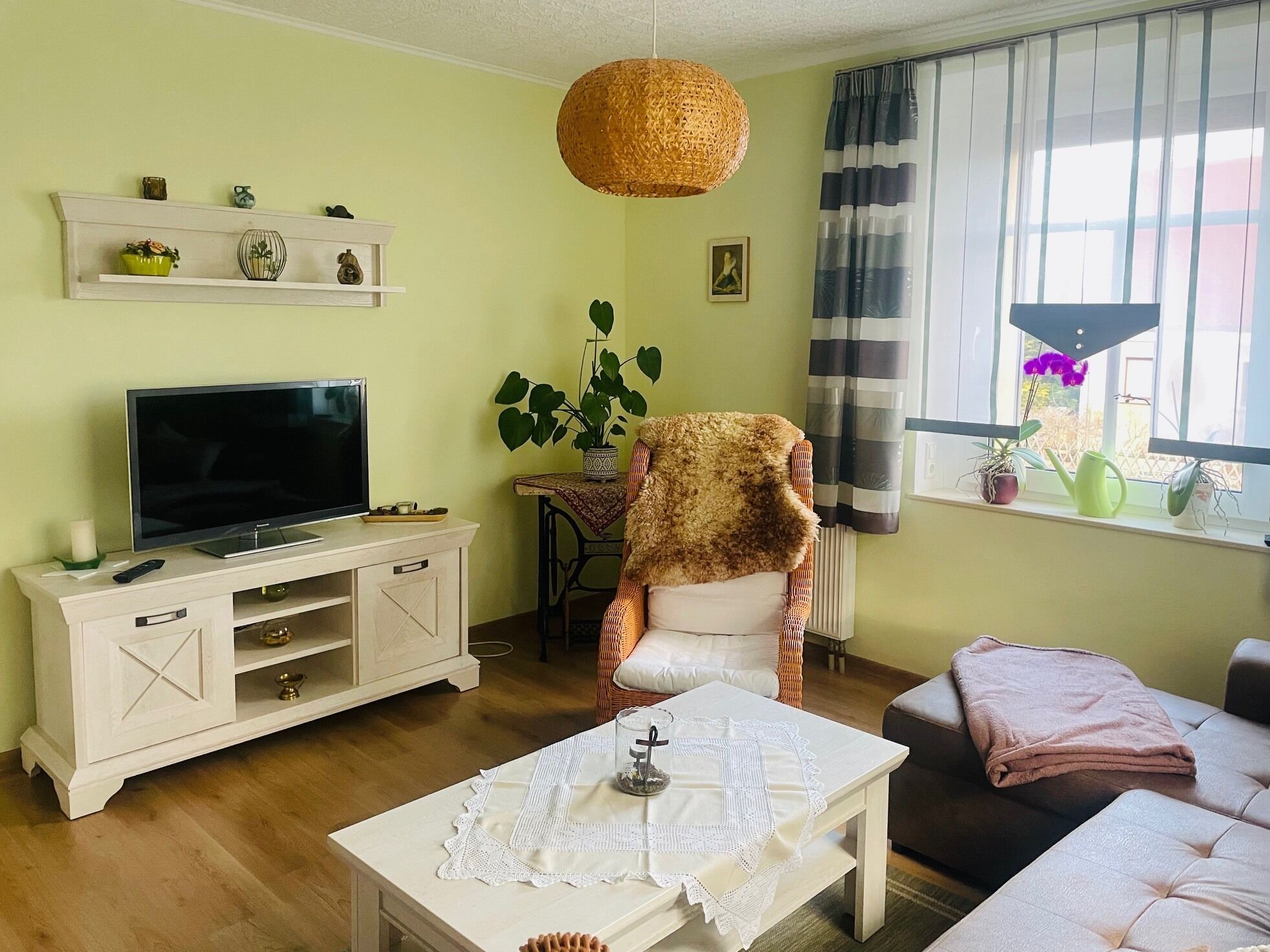 A living room with a TV, white furniture, a wicker hanging light, a plant, and a window with curtains.