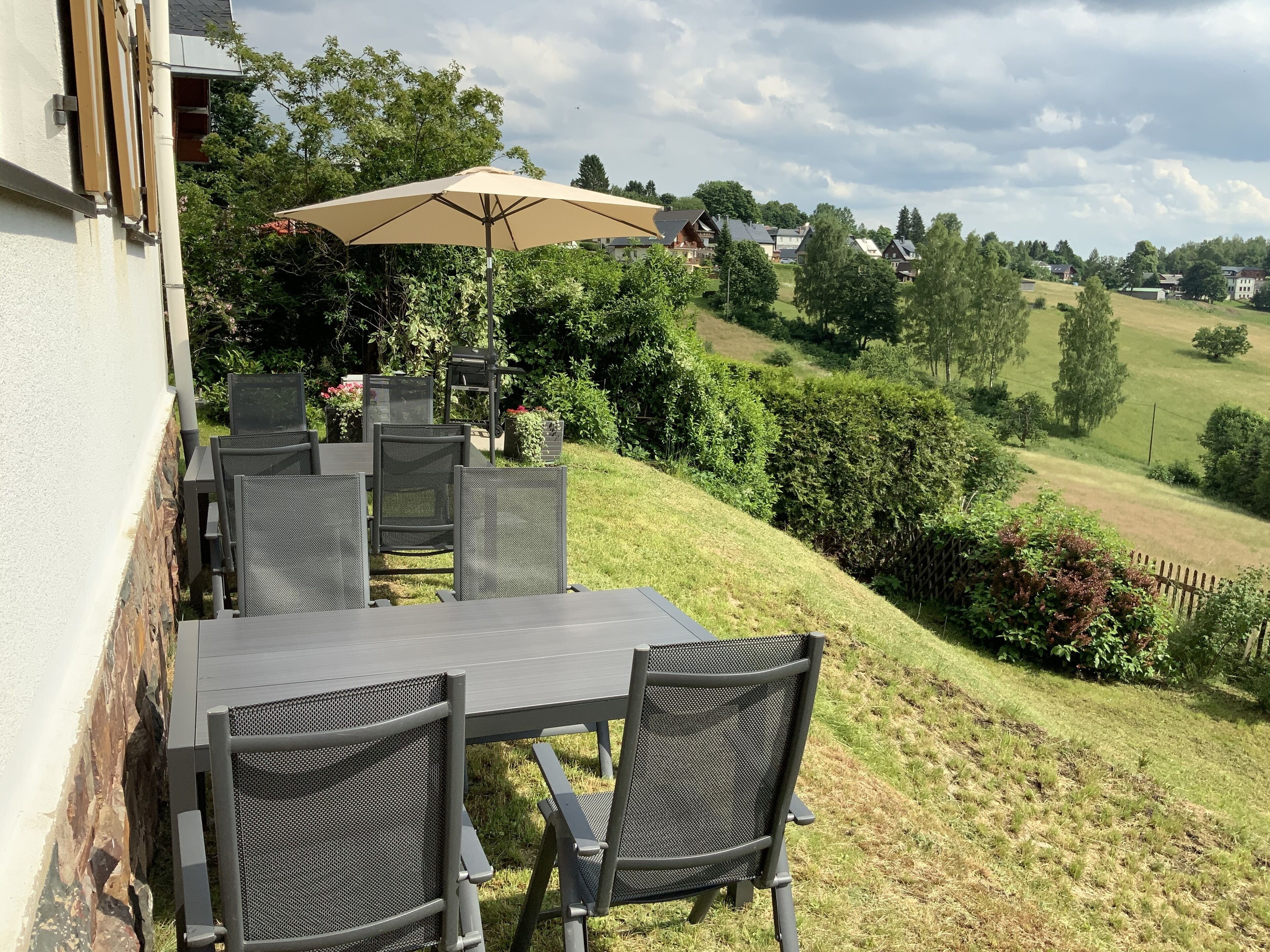 An outdoor patio with a table and chairs, a beige umbrella, and a view of a residential area with houses and trees.