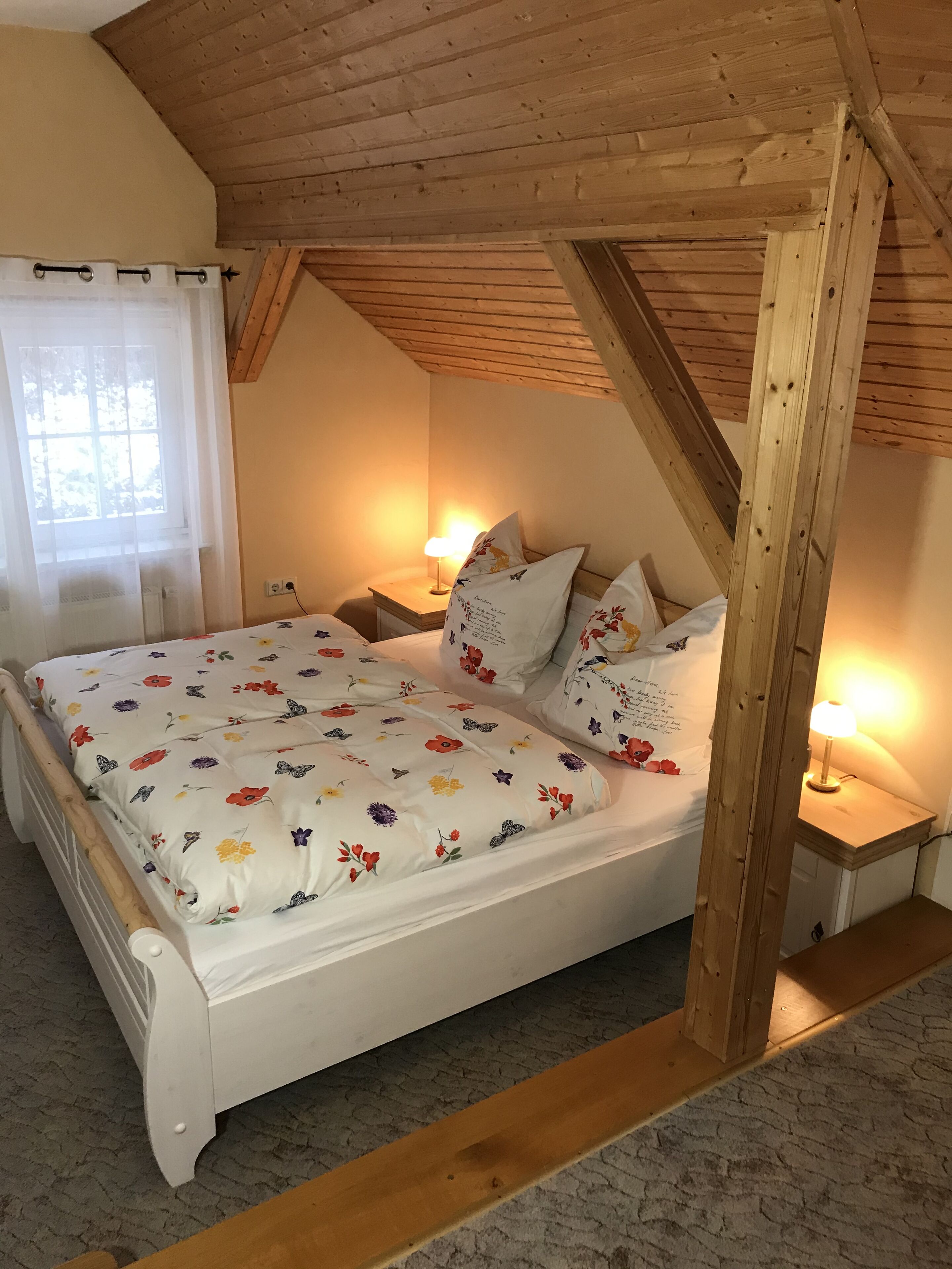 Camera da letto con soffitto in legno, un letto con biancheria da letto floreale, due lampade ai comodini e una finestra con tende leggere.