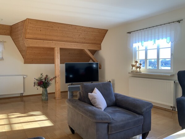 Una sala de estar con techo alto, un televisor de pantalla plana, un sofá, un sillón, una ventana con cortinas y una figurita decorativa de un ciervo.