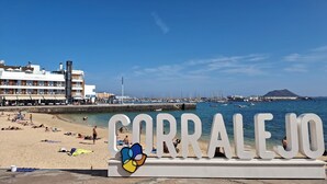 A beach with the sign 'CORRALEJO' and a view of the sea and buildings in the background. - Apartment Gardenia, Oasis Tamarindo, seaview, tropical Garden, 2 Pools (La Oliva)