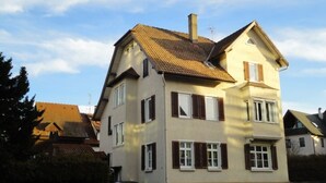 Un bâtiment à deux étages, doté d’un toit en pente, de nombreuses fenêtres avec volets et d’une cheminée bien visible.