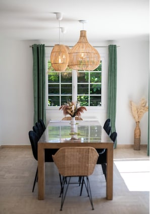 Uma área de refeições com mesa de vidro, cadeiras estofadas pretas e luminárias pendentes de vime.